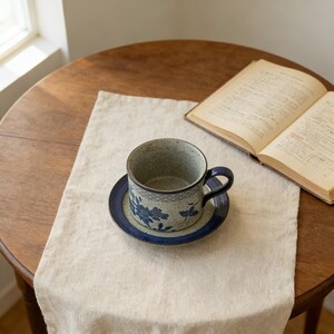 May include: A ceramic teacup and saucer set with a blue floral design. The cup has a dark blue handle and sits on a matching saucer. The set is on a cream-colored linen runner on a wooden table, with an open book in the background.