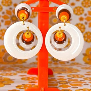Peut inclure: Une paire de boucles d'oreilles rétro avec un schéma de couleurs blanc, orange et marron. Les boucles d'oreilles présentent un design en couches de cercles concentriques et sont suspendues à des crochets dorés. Elles sont présentées sur un support orange.