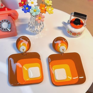 May include: A pair of retro-style earrings with a brown, orange, yellow, and white square design. The earrings have a circular top with matching colors. The earrings are displayed on a white table with other decorative items.