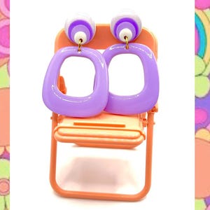 Peut inclure: Une paire de boucles d'oreilles carrées lavande avec des accents blancs et violets, présentées sur une petite chaise orange. Les boucles d'oreilles ont une esthétique rétro et groovy, sur un fond psychédélique coloré.