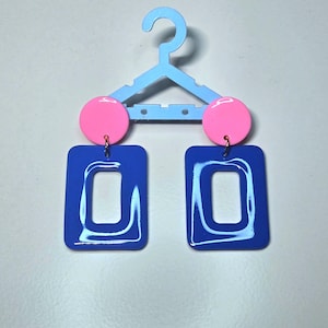 Puede incluir: Un par de pendientes geométricos llamativos. Cada pendiente presenta un círculo rosa brillante conectado a un rectángulo azul vibrante con un centro recortado. Los pendientes están suspendidos de una percha azul claro.