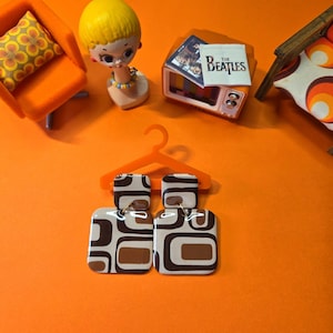 May include: Retro-themed image featuring a miniature orange chair with a patterned pillow, a doll head, a small TV with a Beatles album cover, and a pair of earrings with a brown and white geometric design. The earrings hang from an orange hanger.