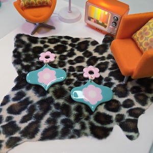Peut inclure: Une paire de boucles d'oreilles en forme de fleur turquoise et rose. Les boucles d'oreilles sont posées sur un tapis à imprimé léopard, avec des chaises oranges et une télévision rétro en arrière-plan. Esthétique rétro et vintage.