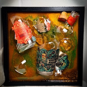 Könnte beinhalten: Ein gerahmtes Kunstwerk mit Glasscherben von Spirituosenflaschen, mit leuchtenden roten, grünen und goldenen Akzenten. Die Etiketten mit Text sind teilweise erhalten, vor einem strukturierten Hintergrund. Der Rahmen ist schwarz.