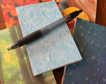 May include: A stack of five small, colorful notebooks with a black pen on top. The notebooks have different designs, including fall leaves, candy corn, a seashell, a blue water pattern, and a blue pattern with circles.