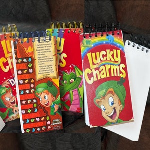May include: Four small spiral notebooks with Lucky Charms cereal box covers. The covers feature the Lucky Charms leprechaun, a dragon, and a game board. The notebooks are blank inside.