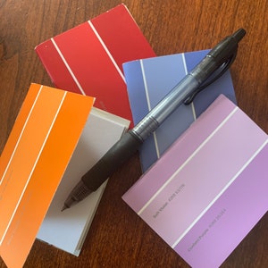 May include: A black pen rests on a collection of paint color swatches. The swatches display shades of red, orange, blue, and purple. The paint swatches are arranged on a wooden surface.