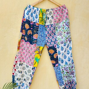 Op de afbeelding: Patchworkbroek met een verscheidenheid aan kleurrijke bloemen- en dierenprints. De broek hangt aan een houten hanger tegen een gele muur. De broek heeft een mix van roze, blauwe, gele en witte stoffen.