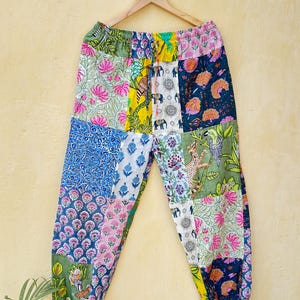 May include: Patchwork trousers featuring a mix of floral and geometric designs in blue, pink, green, and yellow. The trousers are suspended on a wooden hanger against a yellow wall.