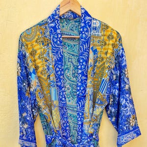 May include: A blue patterned robe with a tie waist. The robe features a mix of floral and paisley designs in shades of blue, yellow, and teal. The robe is hanging on a wooden hanger.