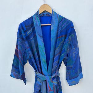 May include: A blue patterned robe with a matching belt hangs on a wooden hanger. The robe features a marbled design in shades of blue, teal, and hints of red and yellow. The robe has three-quarter sleeves and a shawl collar.