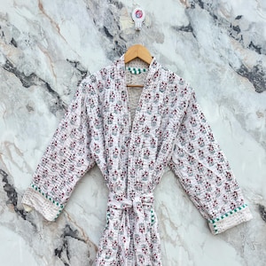 May include: White quilted robe with a floral pattern in red and green. The robe has long sleeves, a tie waist, and green trim along the edges. The robe is hanging on a wooden hanger against a marble background.