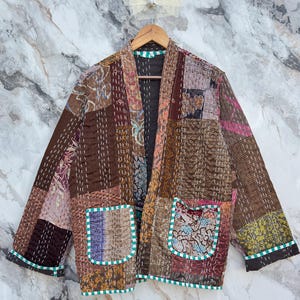 Könnte beinhalten: Patchwork-Jacke in Braun-, Burgunder- und Beigetönen mit einer Vielzahl von Stoffmustern. Die Jacke hat zwei Vordertaschen mit türkisfarbenem und weißem Besatz sowie einen grünen Besatz am Kragen und an den Manschetten. Die Jacke hängt an einem Holzkleiderbügel.