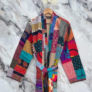 May include: A colorful patchwork robe with long sleeves and a matching belt. The robe is made of various fabric patterns in red, blue, green, yellow, and purple. The robe is hanging on a wooden hanger against a marble background.