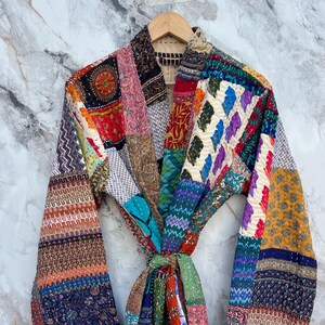 May include: A colorful patchwork robe with a tie closure. The robe features a variety of patterned fabrics in shades of red, blue, green, and yellow. The robe is hanging on a wooden hanger against a marble background.