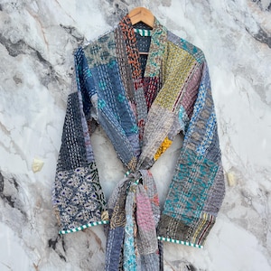 May include: A patchwork robe in various colors, including blue, gray, yellow, and red, hangs against a marble background. The robe features a tie closure and is made of stitched fabric squares. The edges are trimmed with a green and white striped pattern.