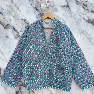 Peut inclure: Veste matelassée verte à motifs floraux rouges et violets. La veste a deux poches avant et est bordée de tissu rayé blanc et vert. La veste est suspendue à un cintre en bois.