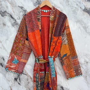 May include: A patchwork robe in shades of orange, red, and brown, with a matching belt. The robe features a variety of patterned fabrics and decorative stitching. The robe is hanging on a wooden hanger against a marble background.