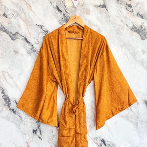 Puede incluir: Una bata de color amarillo mostaza con mangas anchas y acampanadas y un cinturón a juego en la cintura. La prenda tiene un diseño largo abierto por delante con un sutil y fino estampado tono sobre tono en la tela, colgada de una percha.
