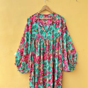 Puede incluir: Un vestido de manga larga con un vibrante estampado floral en tonos rosa brillante, verde azulado y melocotón suave. La prenda presenta una silueta holgada y fluida, un escote en V con finos lazos de cierre y puños fruncidos en las mangas.