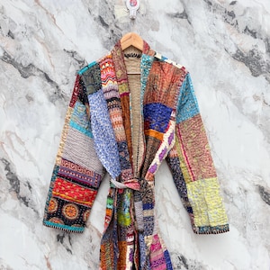 Può includere: Un accappatoio patchwork con cintura annodata, esposto su una gruccia di legno. L'accappatoio presenta una varietà di toppe di tessuto colorate nelle tonalità del rosso, blu, verde e giallo. L'accappatoio è realizzato in tessuto trapuntato.
