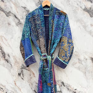 May include: A patchwork robe in shades of blue, green, purple, and brown, with a matching belt. The robe is made of various patterned fabrics, stitched together. The sleeves and edges are trimmed with a white and green border.