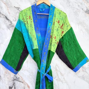 Puede incluir: Una colorida bata tipo kimono de estilo patchwork. La bata presenta paneles de tela verde, azul y negra con estampados florales y de cachemira. Un cinturón azul anuda la cintura. La bata está colgada en una percha de madera contra una pared con efecto mármol.