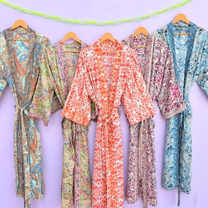 Può includere: Cinque accappatoi colorati con motivi paisley appesi a grucce di legno contro un muro viola. Gli accappatoi sono in tonalità di blu, arancione, viola e verde. Ogni accappatoio ha una cintura abbinata. Una ghirlanda di perline gialle e verdi è appesa sopra.