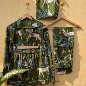 May include: Green pajama set with a matching tote bag, featuring a white leopard and tropical foliage print. The set includes a long-sleeved button-down shirt, shorts, and long pants, all trimmed with white piping. The tote bag hangs on a wooden hanger.