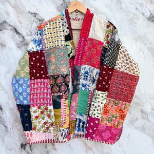 May include: A colourful patchwork jacket made from various fabric squares. The jacket is displayed on a wooden hanger against a marble backdrop. It features floral, geometric, and paisley patterns in its design.