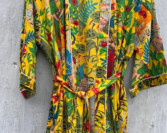 May include: A yellow robe with a vibrant, colorful print featuring figures, foliage, and floral patterns. The robe has white trim and a matching belt. The design includes red, green, blue, and orange hues.