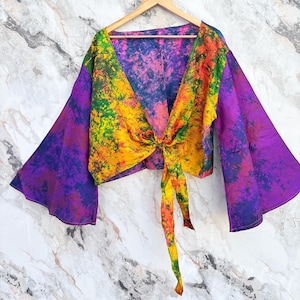Può includere: Top con nodo frontale e vivace fantasia tie-dye nei toni del viola, giallo, verde e arancione. Il top presenta ampie maniche a campana ed è esposto su una gruccia di legno su sfondo marmoreo.