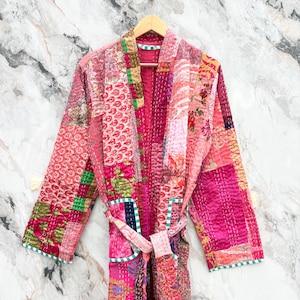 May include: A patchwork robe in shades of pink, red, and green, with a matching belt. The robe is made of various fabric patterns and textures, and is hanging on a wooden hanger against a marble background.