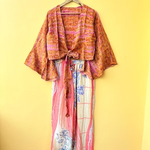 Könnte beinhalten: Ein zweiteiliges Outfit bestehend aus einem bauchfreien Wickeloberteil mit weiten Glockenärmeln und einem passenden langen Rock. Das Oberteil hat ein orange-rosa gemustertes Gewebe mit Bindegürtel. Der Rock zeigt ein florales Muster in Creme, Rosa und Blau.