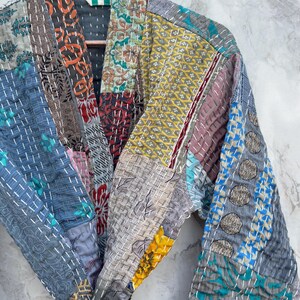 May include: A patchwork kimono-style robe in various colors, including blue, gray, yellow, and red. The robe is made of multiple fabric squares stitched together with white thread. The garment is hanging on a wooden hanger against a light gray background.