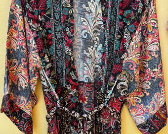 May include: A long, open-front robe with a vibrant floral and paisley pattern in red, teal, and yellow on a black background. The robe has long sleeves and a matching tie belt. The garment is hanging on a wooden hanger.