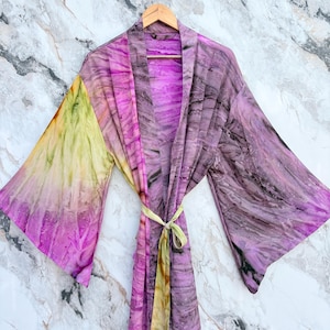 May include: A tie-dye kimono robe in shades of purple, pink, yellow, and green hangs on a wooden hanger. The robe has wide sleeves and a matching tie belt. The fabric has a marbled effect.
