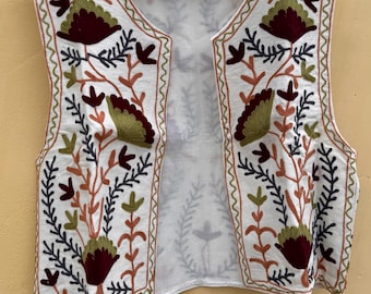 May include: White embroidered vest with a floral design in shades of green, burgundy, and orange. The vest is sleeveless and has a zigzag stitch trim. It hangs on a wooden hanger against a yellow background.