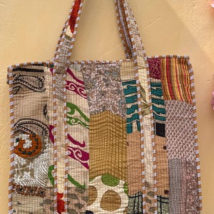 Może przedstawiać: Torba typu tote z patchworku z różnymi wzorami i kolorami tkanin, w tym paisley, paski i kropki. Torba ma prostokątny kształt z pasującym uchwytem i wykończeniem. Torba wykonana jest z pikowanej tkaniny.