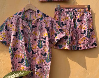 May include: Pink pajama set with a bird and floral pattern. The set includes a short-sleeved button-down shirt, shorts, and a matching drawstring bag. The design features black birds, yellow accents, and blue and green foliage.