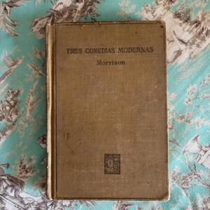 May include: A vintage book titled "Tres Comedias Modernas" by Morrison. The book has a brown cover with a small square design on the front.