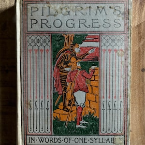 May include: A vintage book titled "Pilgrim's Progress" in words of one syllable. The cover features an illustration of two figures in a landscape setting. One figure is wearing armor and pointing towards a stone wall, while the other figure is holding a staff and looking at the wall.