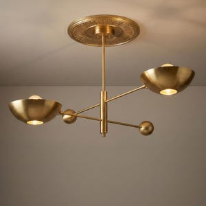 May include: A brass-toned chandelier with three bowl-shaped lights. The fixture is attached to the ceiling with a decorative circular plate. The lights are on, emitting a warm glow. A modern lighting fixture, suitable for a living room or dining area.