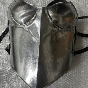 May include: A polished silver-coloured metal breastplate featuring a central vertical ridge and moulded chest contours. The surface displays a subtle, etched crackle pattern throughout. Black leather straps with metal buckles are attached to the sides..