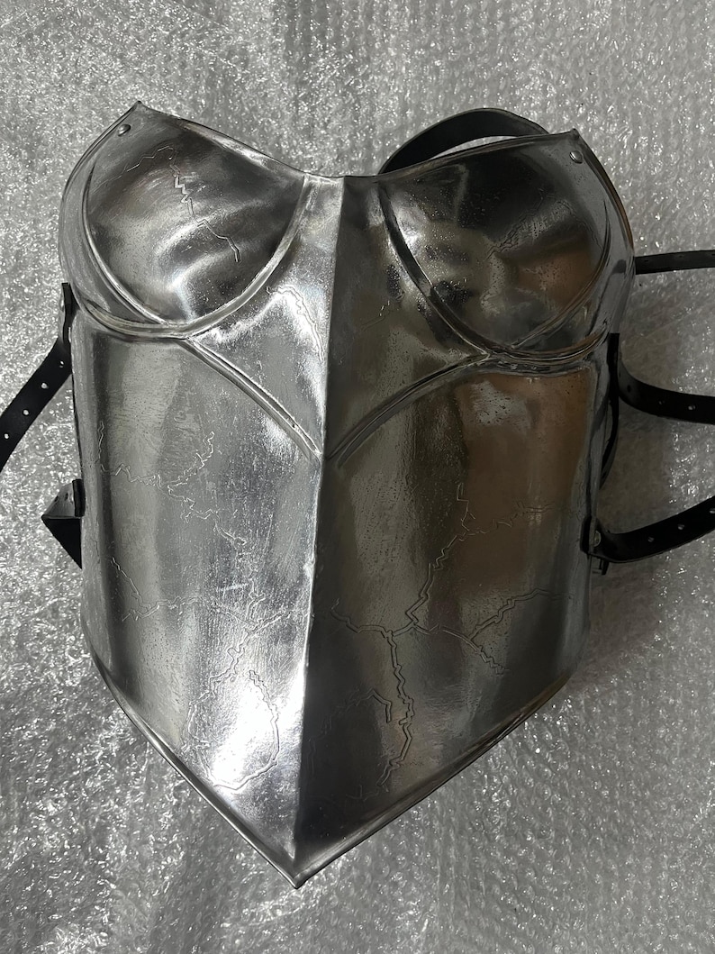 May include: A polished silver-coloured metal breastplate featuring a central vertical ridge and moulded chest contours. The surface displays a subtle, etched crackle pattern throughout. Black leather straps with metal buckles are attached to the sides..