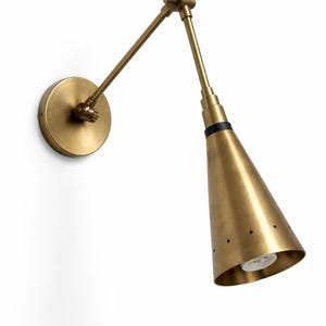 May include: A brass wall lamp with an adjustable arm and a conical shade. The lamp features a circular base and a black band near the top of the shade. The shade has small, evenly spaced holes. The lamp is designed for wall mounting.