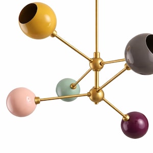 Op de afbeelding: Een moderne kroonluchter met een gouden afwerking en zes kleurrijke bollen. De bollen zijn in tinten geel, roze, groen, grijs en paars. De lamp heeft een centrale naaf met armen die zich naar elke bol uitstrekken.