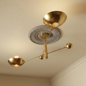 Op de afbeelding: Een moderne, goudkleurige plafondlamp met drie komvormige kappen en bolvormige accenten. De armatuur is gemonteerd op een decoratief plafondmedaillon. Het licht is aan en verlicht de kamer.