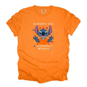 May include: An orange t-shirt with a cartoon character in the centre. The character has blue fur and large ears, surrounded by colourful handprints. Text reads "HARMONY DAY EVERYONE BELONGS".