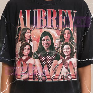 May include: Black t-shirt with a retro graphic of Aubrey Plaza and the text "AUBREY PLAZA". The design includes the Milkshake logo and lightning bolt accents. The shirt has a vintage aesthetic.